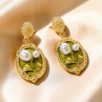 A pair of gold drop earrings with textured gold posts and oval green enamel pendants featuring white faux pearls and gold leaf details, displayed on a beige satin fabric background.