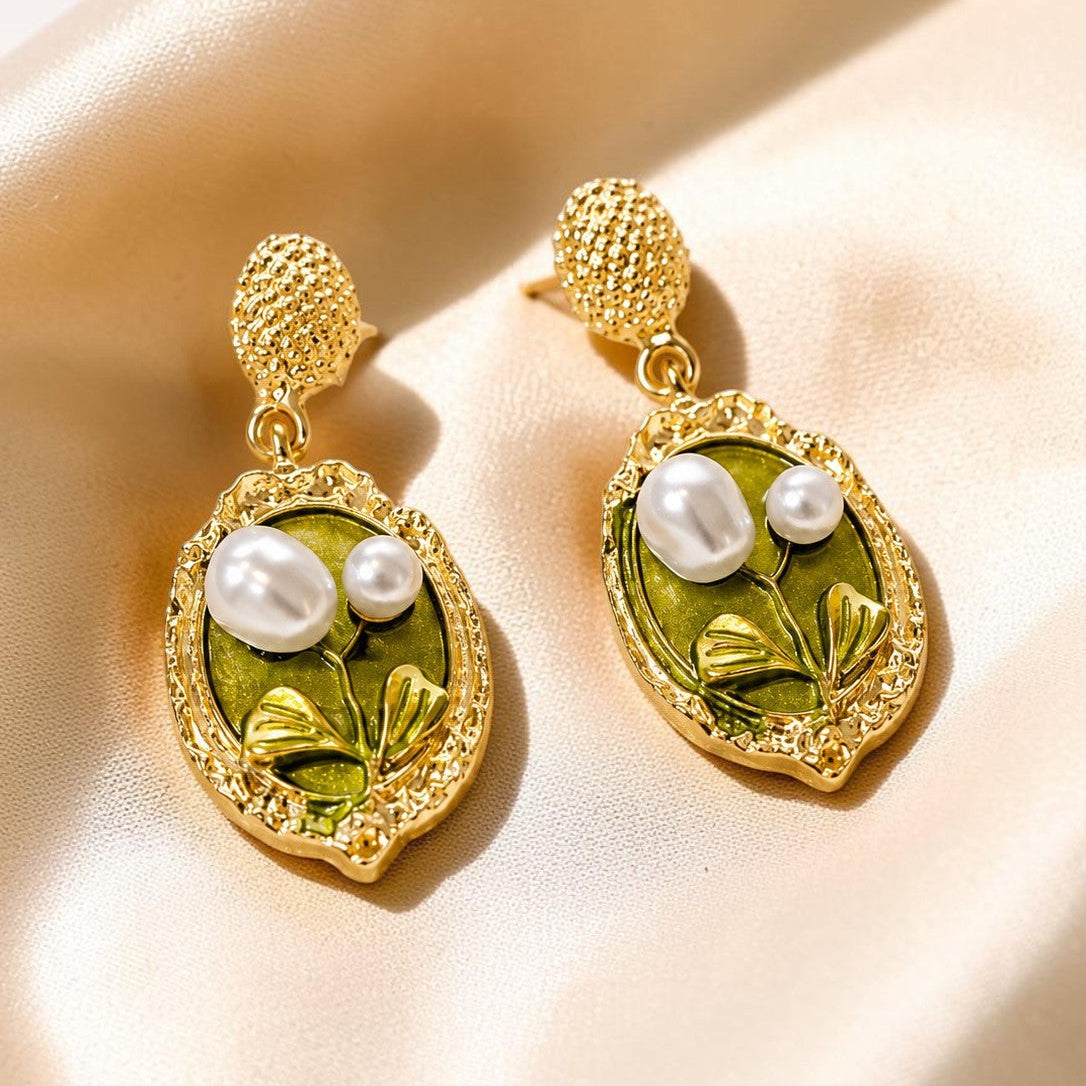 A pair of gold drop earrings with textured gold posts and oval green enamel pendants featuring white faux pearls and gold leaf details, displayed on a beige satin fabric background.