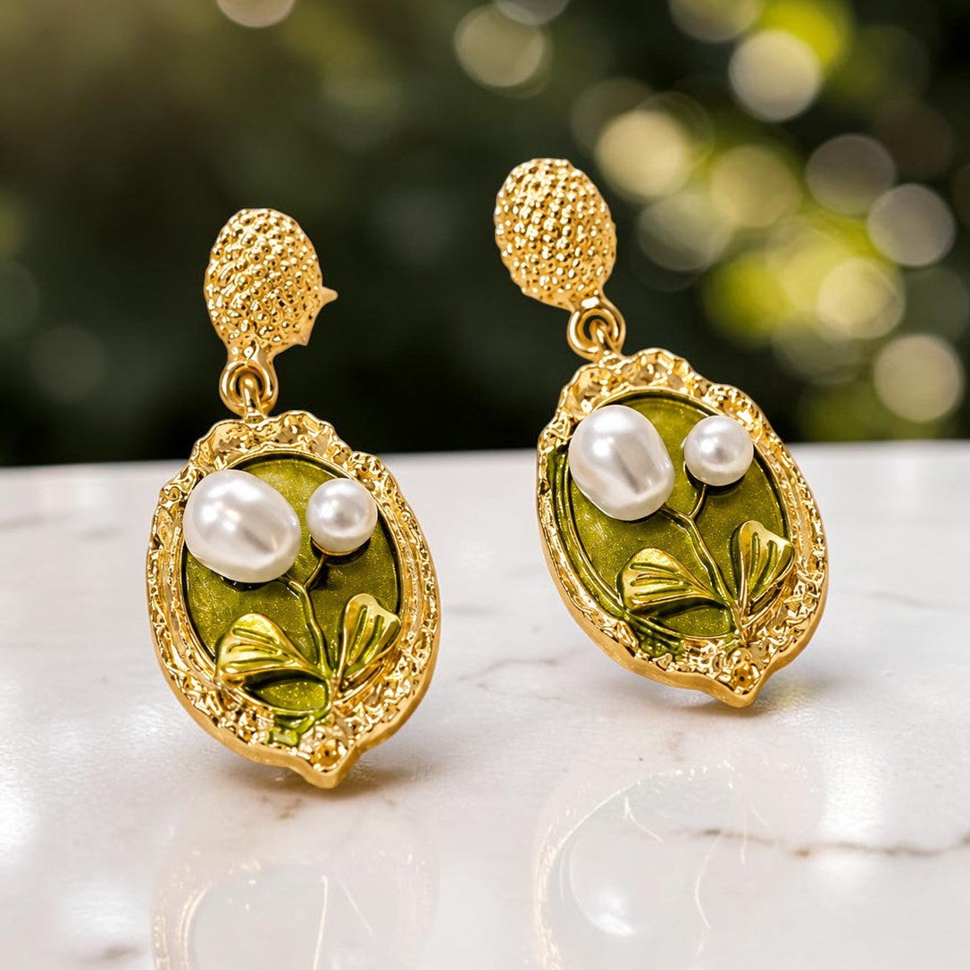 A pair of gold and green drop earrings with white faux pearls, displayed on a white marble surface with a soft green bokeh background.