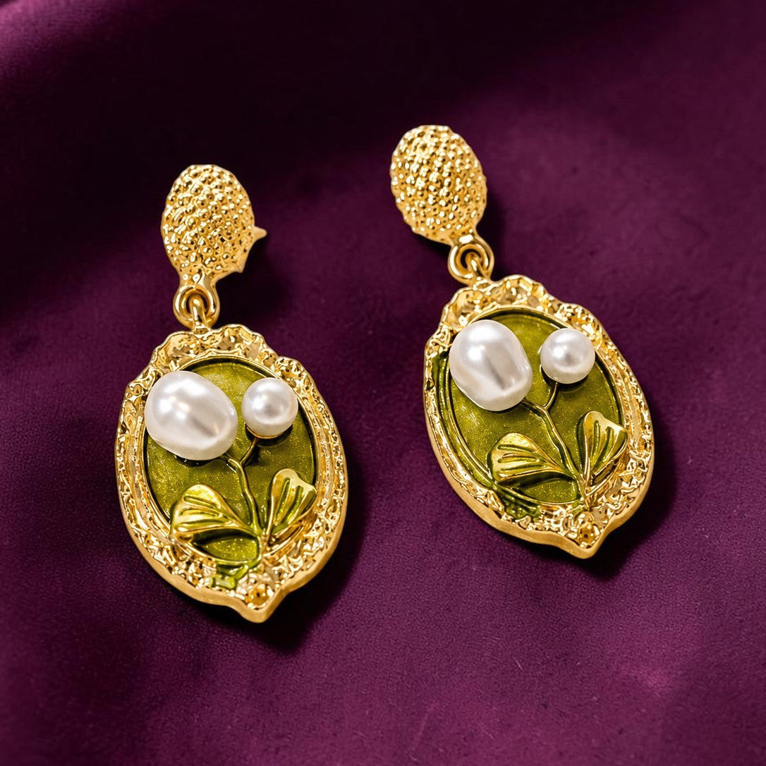 A pair of gold drop earrings with textured top studs and oval leaf-shaped charms featuring green enamel, pearl-like white beads, and small clear rhinestones, displayed on a deep purple satin fabric.
