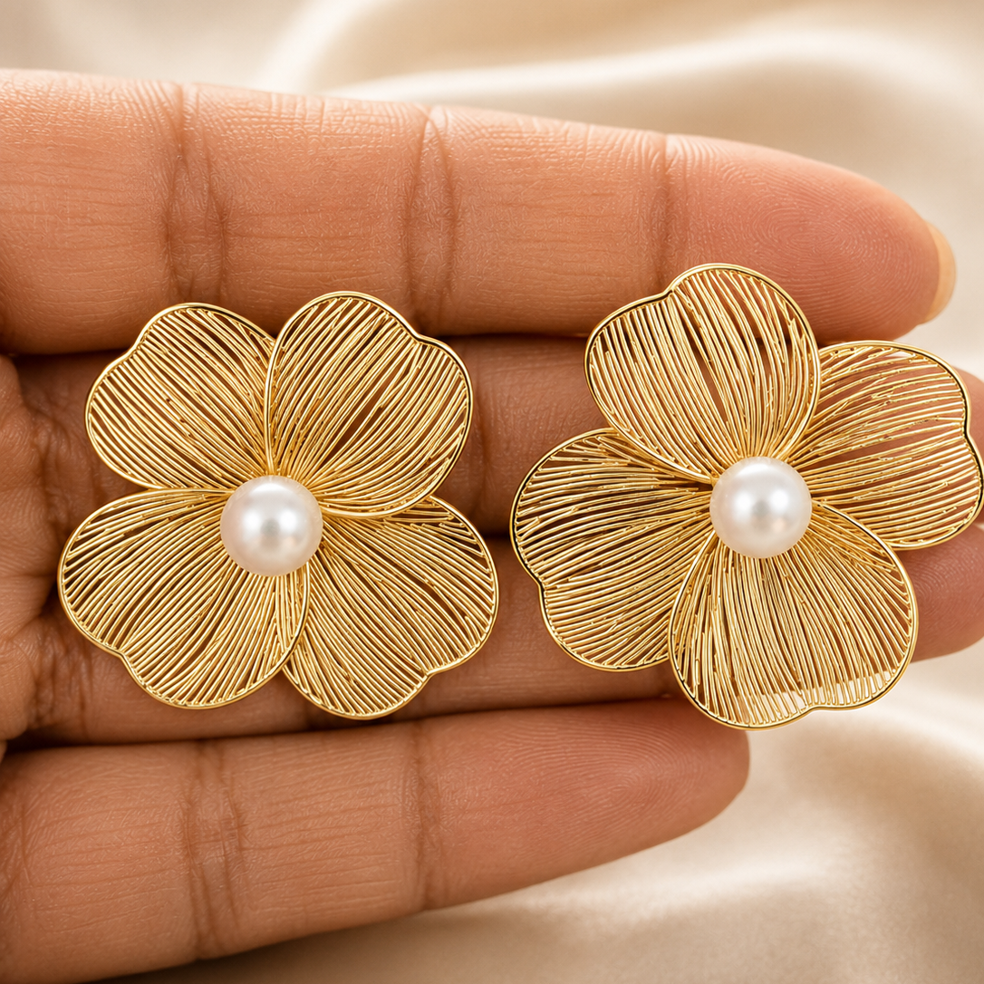 Gold flower-shaped earrings with pearl centers held between fingers on a beige fabric background