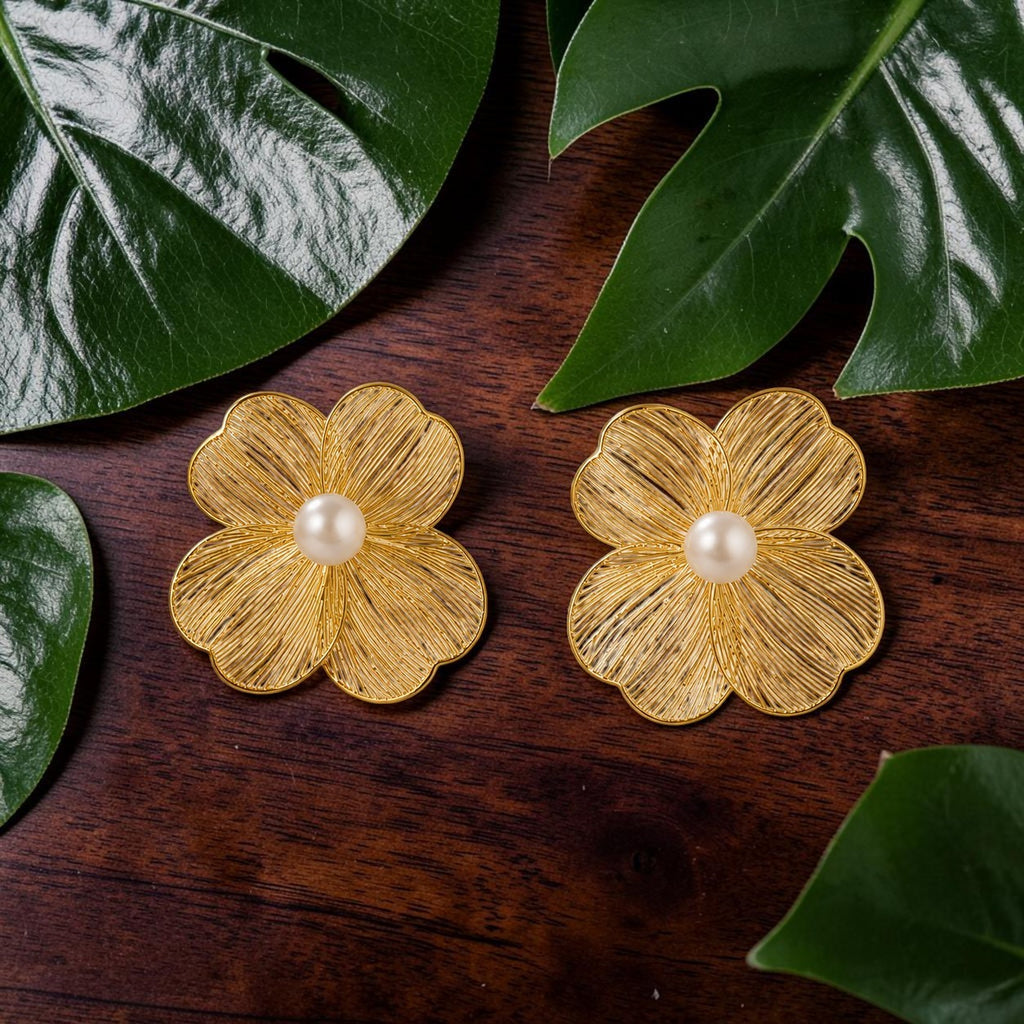 Goldne flower-shaped earrings with a pearl center on a wooden surface surrounded by green leaves.