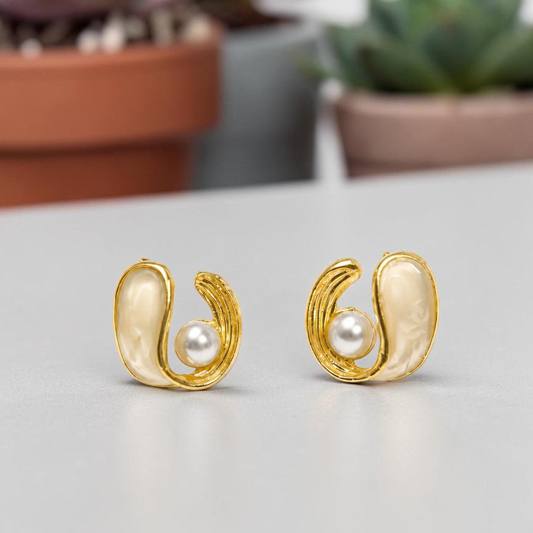 Gold tone earrings with pearls on a light surface, with plants in the background