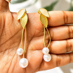Golden leaf earrings with pearl drops held in a hand against a blurred natural background