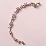 A silver-tone tennis bracelet featuring a line of pink gemstones of various shapes, displayed on a solid pale pink background.