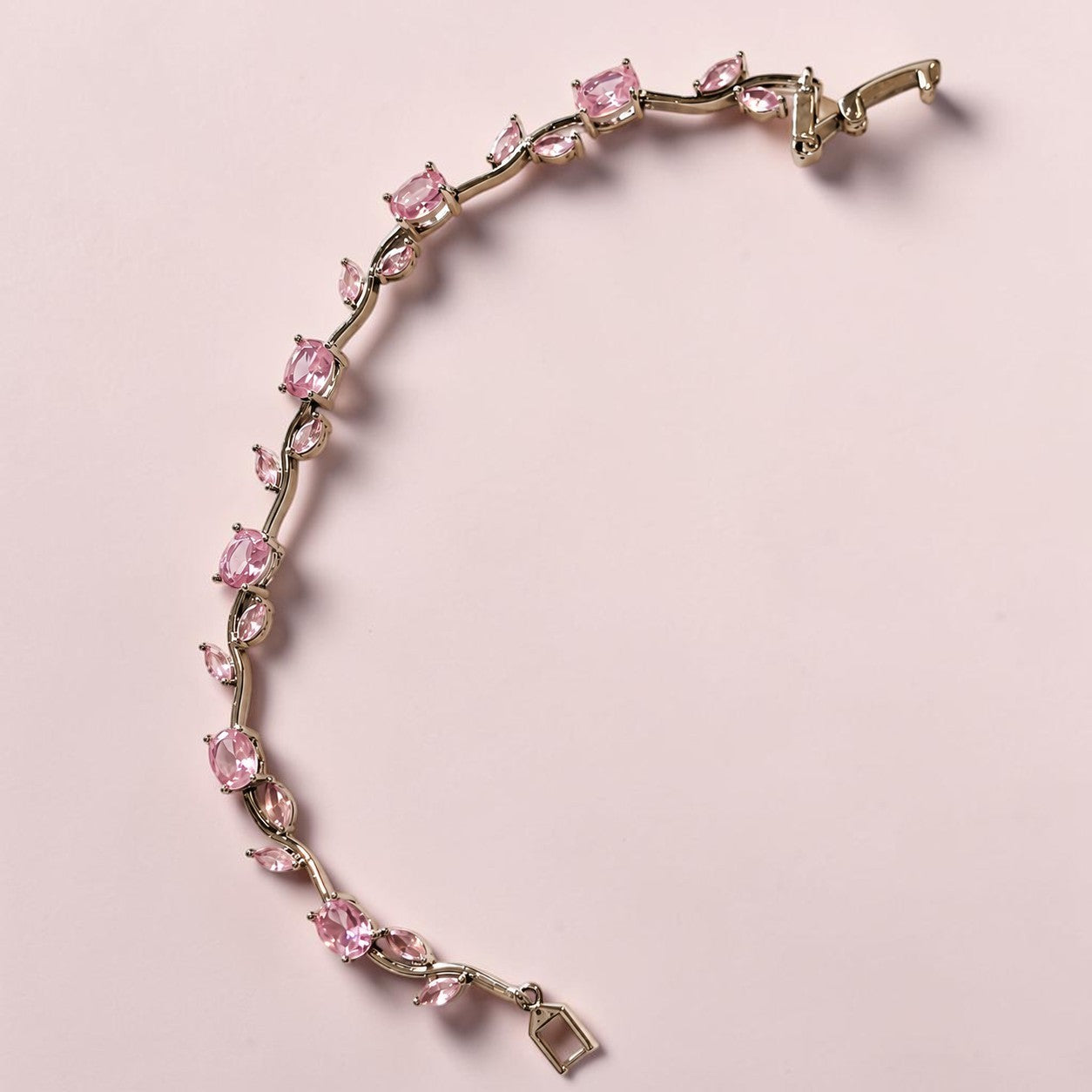 A silver-tone tennis bracelet featuring a line of pink gemstones of various shapes, displayed on a solid pale pink background.