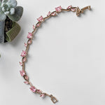 A silver tone bracelet with a series of pink gemstones set along a curved, leaf-like path, displayed on a light gray surface next to a small potted succulent.