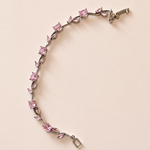 A silver-toned bracelet featuring a vine-like design set with pink gemstones, photographed on a solid pale pink background.