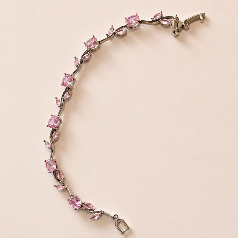 A silver-toned bracelet featuring a vine-like design set with pink gemstones, photographed on a solid pale pink background.