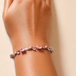 A close-up of a person's hand and wrist wearing a delicate bracelet with pink gemstones and silver-toned metal links, set against a solid beige background.