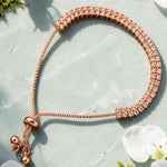 A rose gold adjustable bracelet with a textured, diamond-like paved band and a sliding bead closure, styled on a light gray stone surface with white cherry blossoms and green moss.