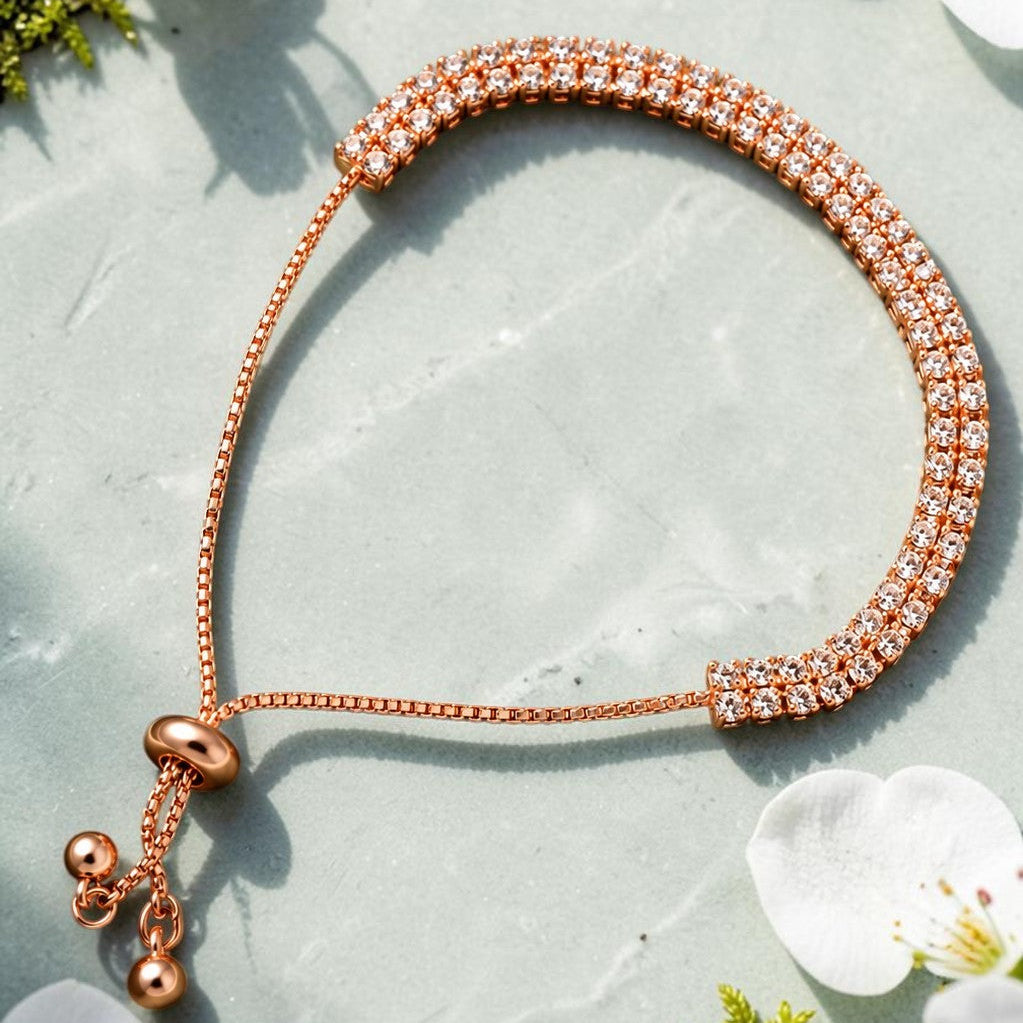 A rose gold adjustable bracelet with a textured, diamond-like paved band and a sliding bead closure, styled on a light gray stone surface with white cherry blossoms and green moss.