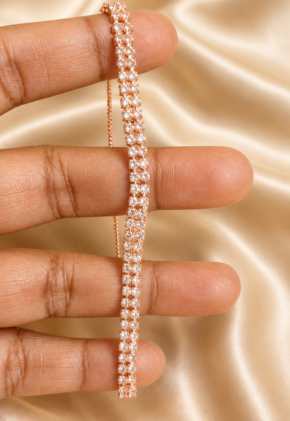 A close-up of a hand holding a delicate rose gold tennis bracelet against a draped champagne satin background.