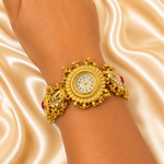 An ornate gold-toned wristwatch with a decorative open bracelet and small pink gemstone accents, shown worn on a person's wrist against a draped satin fabric background.