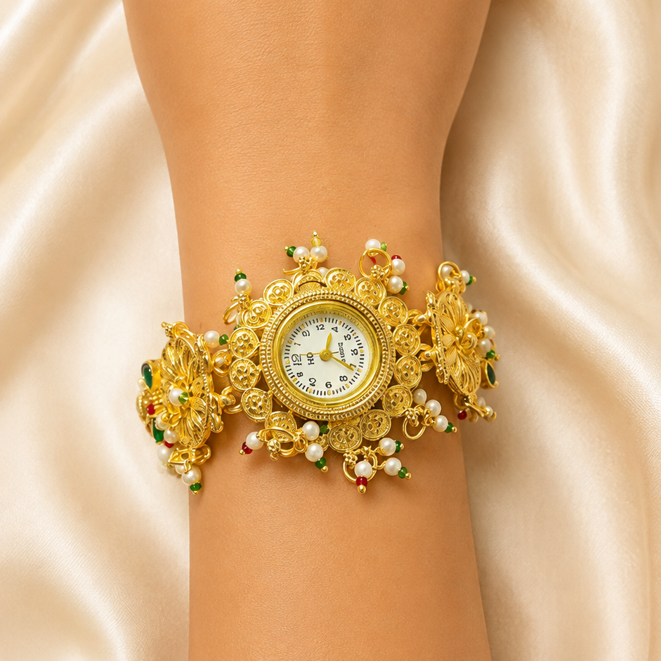 A close-up of a person's wrist wearing an ornate gold cuff watch with white pearl-like beads, green and red jewel accents, and a round white dial, set against a draped champagne satin background.