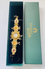 A gold-toned ornate watch with a circular dial and decorative floral details, displayed inside an open teal jewelry box next to its closed box, which has the brand name 'SHINE & RISE' printed in gold.