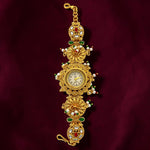 An ornate gold-plated watch bracelet featuring a central round clock face, decorated with green and red enamel, white pearls, and clear stones, set against a dark burgundy fabric background.