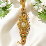 An ornate gold-plated watch with intricate filigree detailing, set with red and green enamel accents and small white stones. The watch is displayed on a draped cream fabric background decorated with green leaves and white flowers.