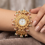 A close-up of a gold-toned ornate wristwatch with pearl and red-green bead accents, worn on a person's wrist against a soft neutral background.