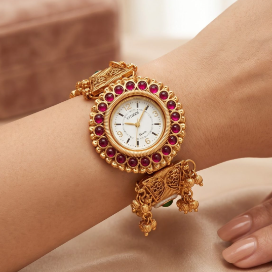A gold-plated bangle watch with a white dial, surrounded by a halo of red gemstones, worn on a woman's wrist against a soft beige satin background.
