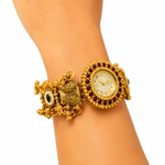 An ornate gold-toned cuff watch bracelet with a round dial, surrounded by dark red stones, and additional decorative elements including a bow charm and green stone accent, shown worn on a wrist against a white background.