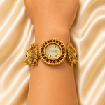 A gold-toned traditional-style wristwatch with a round dial, surrounded by red gemstone accents and decorative side elements, shown worn on a person's wrist against a satin fabric background.
