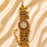 An ornate gold-plated bracelet watch with red and green stone accents, displayed on a draped beige satin fabric background.