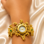 A close-up of a person's wrist wearing an ornate gold cuff watch with floral details and small green stone accents, set against a draped cream satin fabric background.