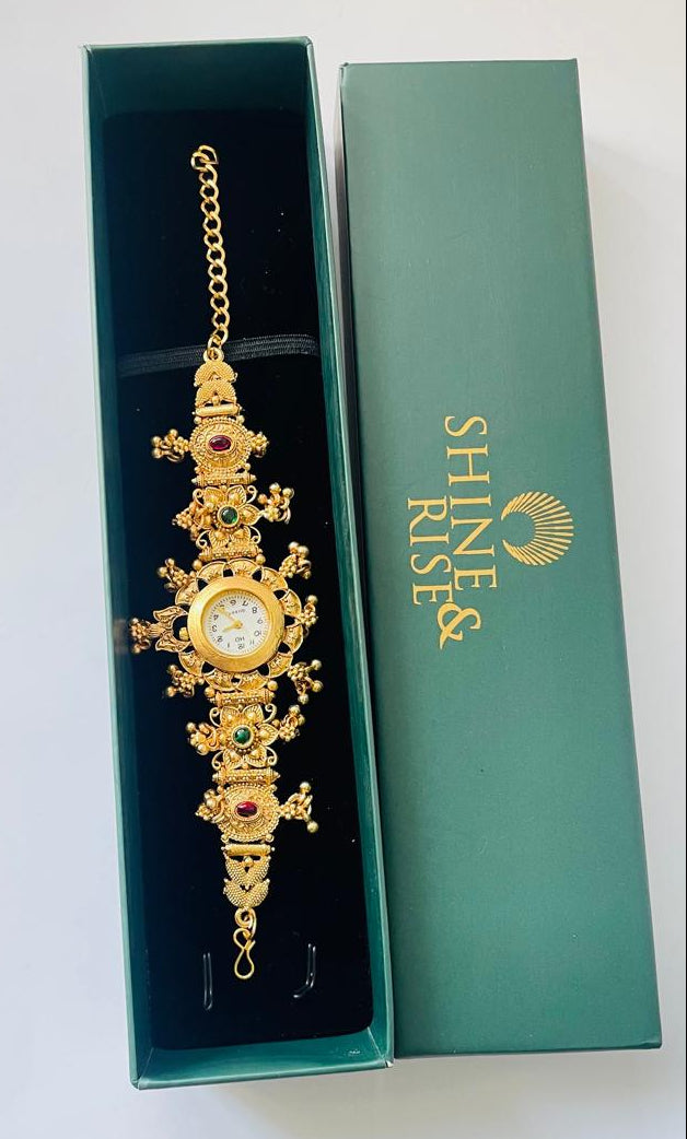 An ornate gold-toned watch bracelet with a central round dial and small green and red gemstone accents, displayed inside an open teal jewelry box next to its branded green lid. The lid shows the brand name Shine Rise.