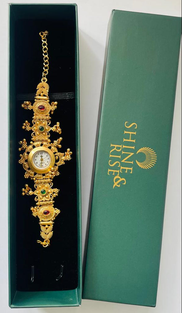 An ornate gold-toned vintage-style wrist watch with green and red gemstone accents, presented inside an open black gift box next to a green box lid printed with the brand name 'SHINE & RISE'.