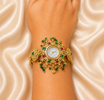 A close-up of a person's wrist and hand wearing an ornate gold watch with green and red gemstone embellishments, set against a draped beige satin fabric background.