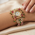 An ornate gold wristwatch with red and green gemstone accents and a pearl-embellished band, shown worn on a person's wrist with a hand resting nearby against a soft beige satin background.