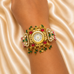 An ornate gold-toned cuff watch with red and green gemstone accents and white crystal-like stones, shown worn on a person's wrist against a draped champagne satin background.