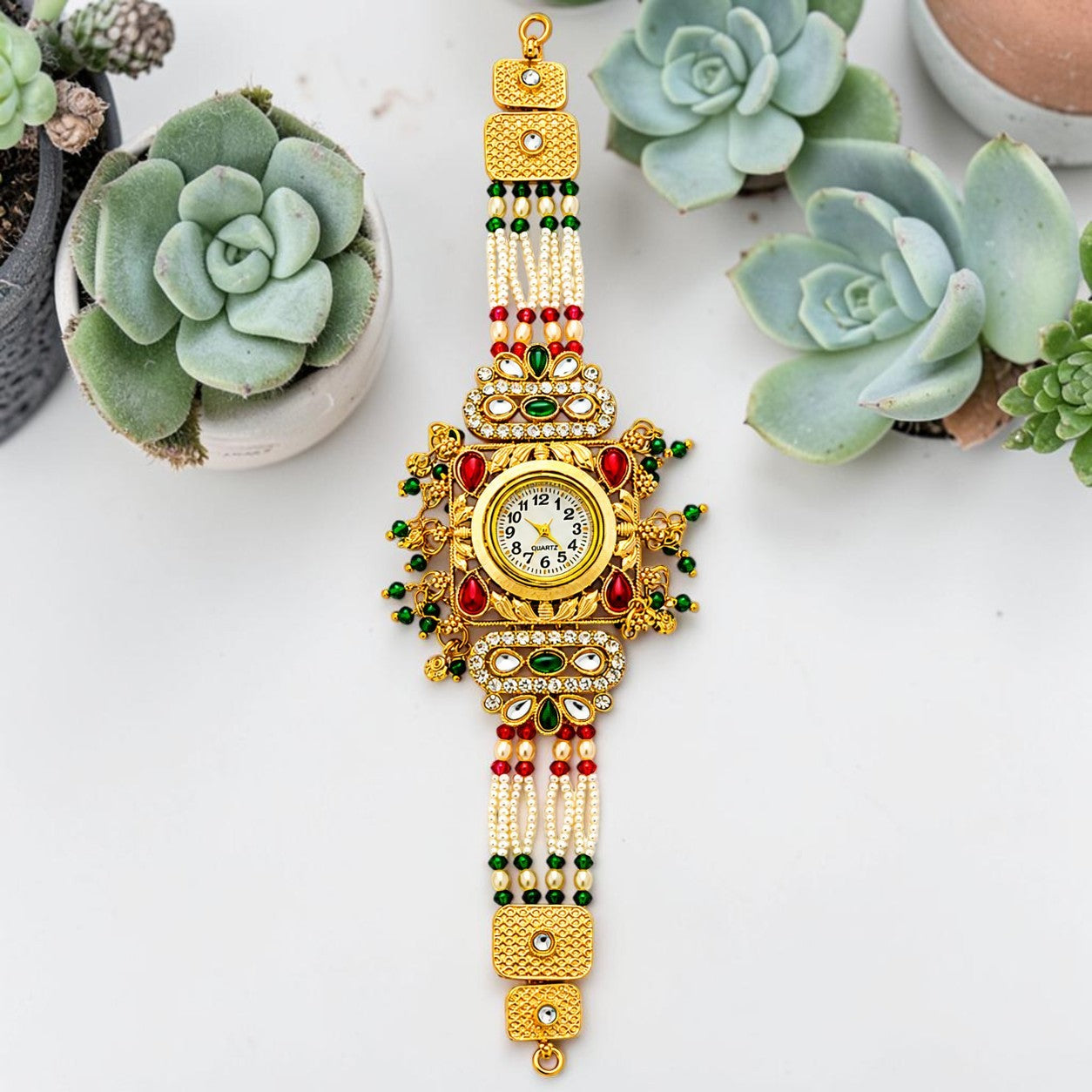 An ornate gold-plated watch with intricate red and green beadwork, white pearl-like beads, and rhinestone embellishments, displayed on a white surface with decorative succulent plants.