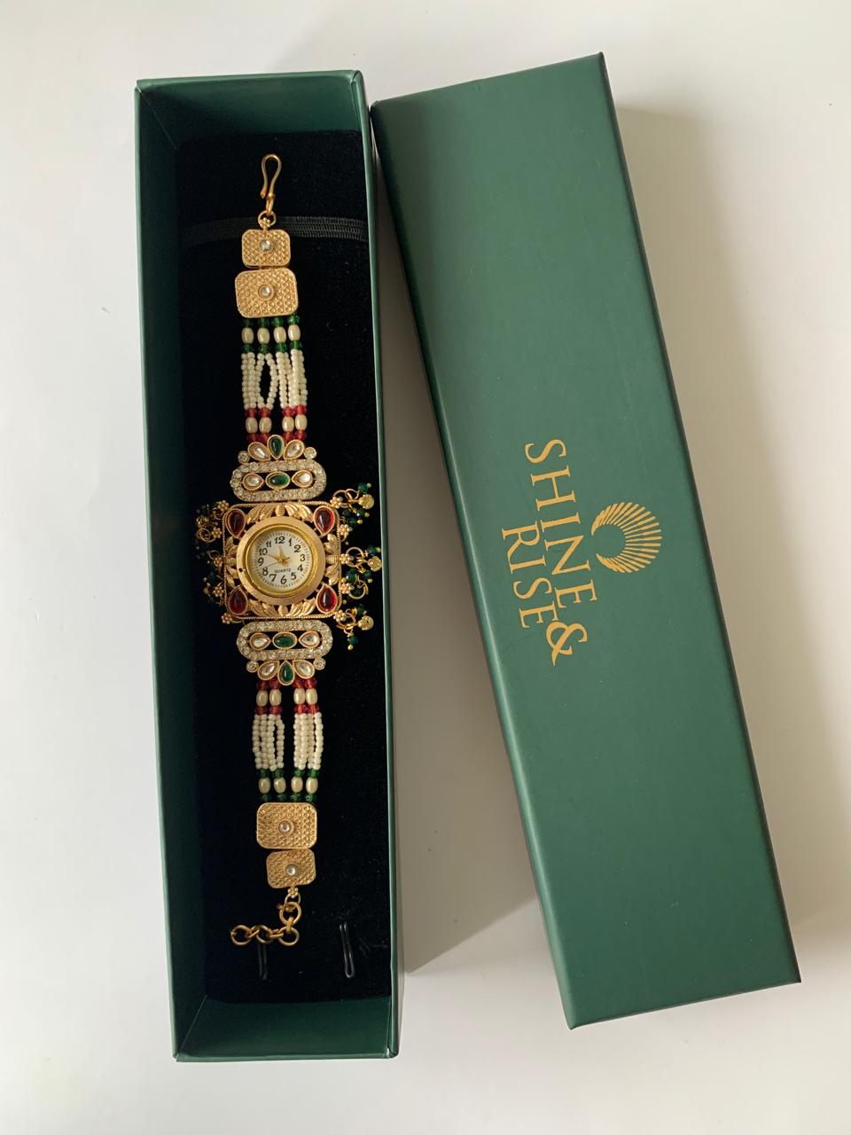 A gold-plated traditional Indian watch bracelet with intricate beadwork and a central watch face, presented inside an open dark green box next to its closed lid which features the brand name 'SHINE RISE' in gold.