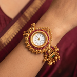 A close-up of a woman's wrist wearing an ornate gold-toned traditional Indian watch with red gemstone accents and dangling bell charms, set against a maroon sari.