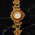 An ornate gold-plated traditional wristwatch with intricate filigree details, green stone accents, and a central round dial, displayed on a black marble surface.