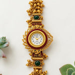 An ornate gold-toned traditional wristwatch with intricate filigree details, green stone accents, and a white dial, displayed vertically on a light background with small succulent plants at the corners.