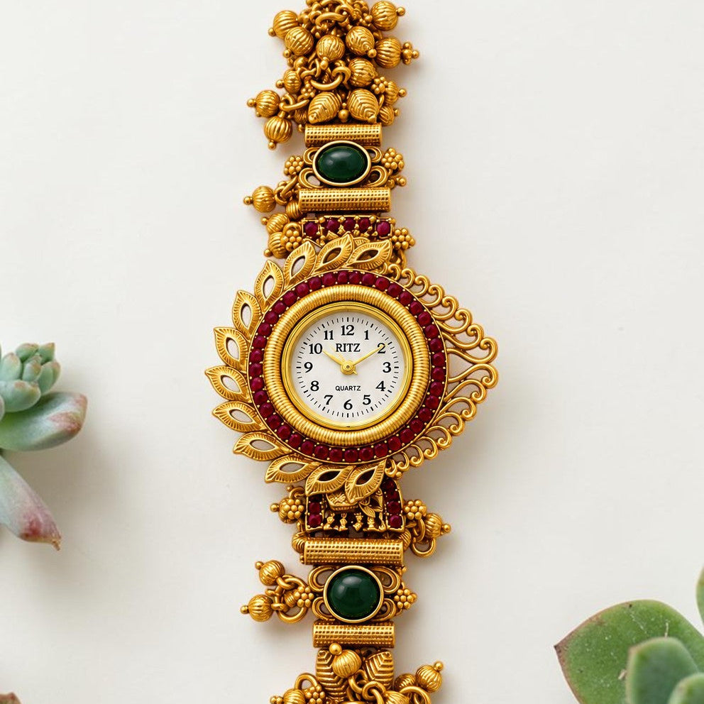 An ornate gold-toned traditional wristwatch with intricate filigree details, green stone accents, and a white dial, displayed vertically on a light background with small succulent plants at the corners.