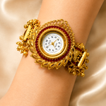A close-up of a gold-toned ornate bracelet watch with a white dial and red gemstone accents, worn on a person's wrist against a satin fabric background.