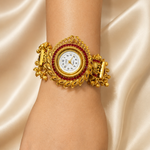 A close-up of a person's wrist wearing an ornate gold cuff watch with a white dial and a red gemstone border, set against a draped champagne satin background.