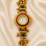 An ornate gold-toned jewelry watch with intricate filigree details, green stone accents, and a white dial, displayed on a draped beige satin fabric background.