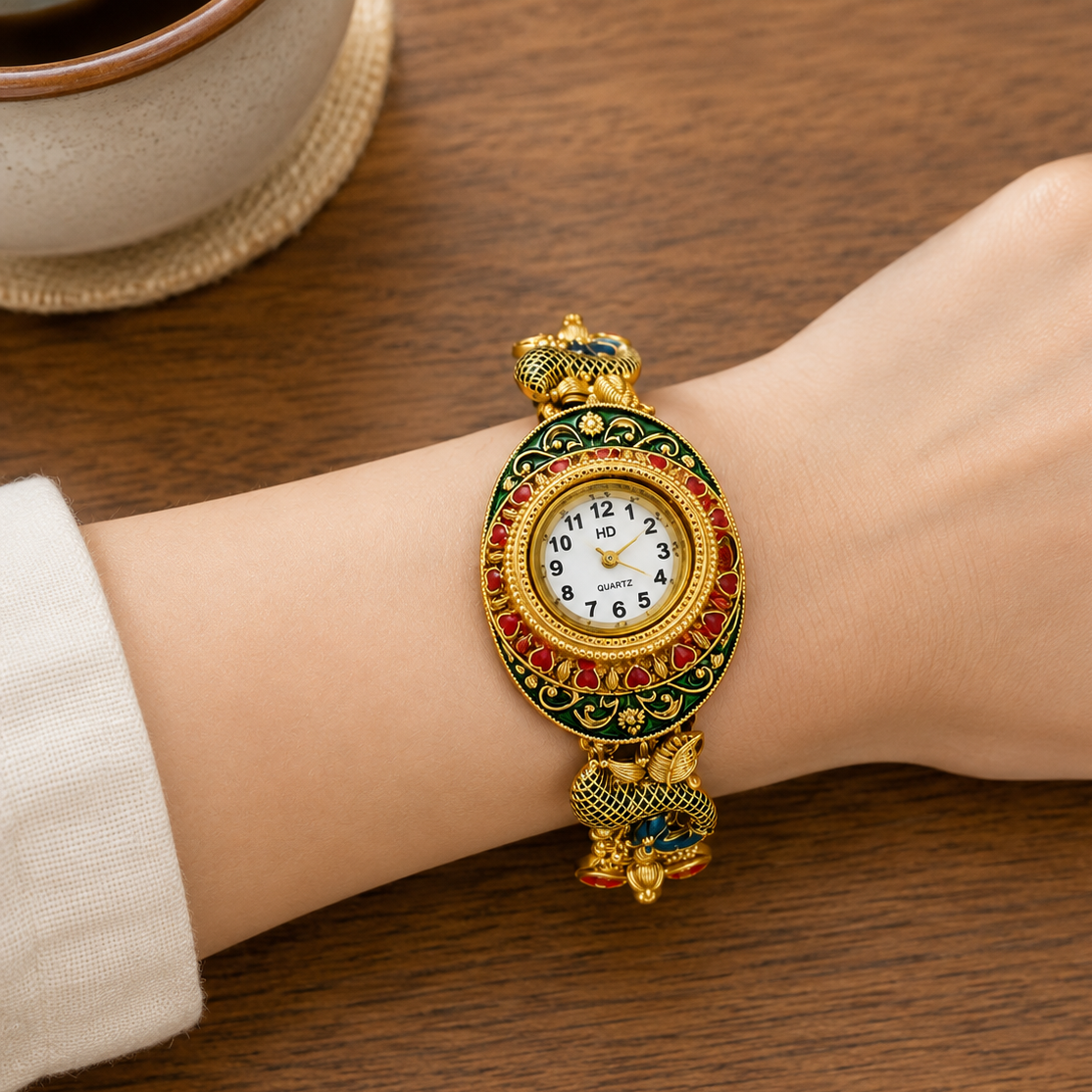 A close-up of a person's wrist wearing a decorative gold-tone watch with red and green enamel detailing, a white dial, and a matching bracelet, resting on a wooden table.