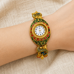 A gold-tone watch with a snake-shaped bracelet and colorful green, red, and blue enamel details, shown worn on a person's wrist against a beige fabric background.