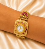 A close-up of a person's wrist wearing a traditional Indian gold-plated watch with a decorative dial, pink stone accents, and a peacock motif on the band, set against a draped champagne satin background.