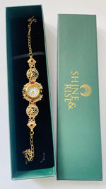 A gold-toned ornate watch bracelet with red and white gemstone accents, displayed inside a dark green presentation box next to its matching green box lid featuring the brand name 'SHINE & RISE'.