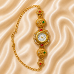 An ornate gold chain watch with decorative filigree details, red and green gemstones, and a round white dial, displayed on a beige satin fabric background.