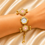A gold-plated women's watch with pink gemstone accents and crystal details, shown on a model's wrist against a gold satin background.