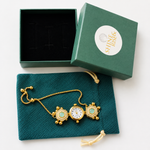 Gold-tone pocket watch necklace with turquoise inlays, displayed on a dark teal drawstring pouch next to its open presentation box and branded lid. The box lid features a white circular logo reading 'SHINK & RICE'.