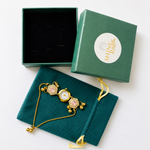 A gold-tone watch with pink crystal accents and a white dial, displayed on a dark teal velvet jewelry pouch next to its green presentation box and inner box lid. The brand name 'SHINE KINGS' is visible on the outer box lid.